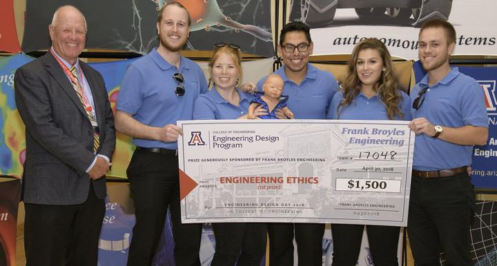 A man in a suit next to five students wearing blue polo shirts and holding a giant check for $1,500