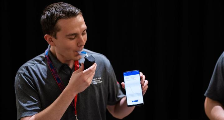 A student wearing a button up shirt holds a phone while demonstrating how to use a breathing device.