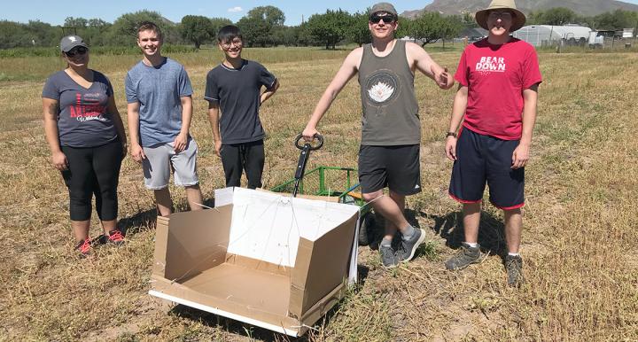 Five people standing in a field with a box attached to a cart