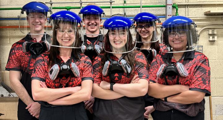 Six students pose indoors wearing welding helmes