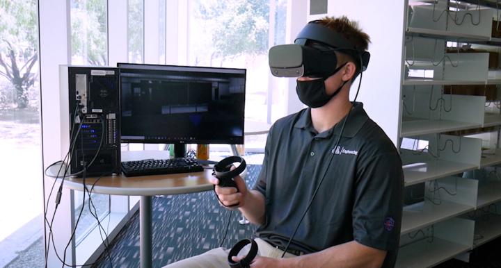 A student wearing a virtual reality headset and sitting next to a computer monitor.