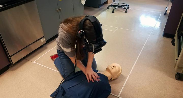 A woman wearing a virtual reality headset kneeling over a CPR practice dummy.