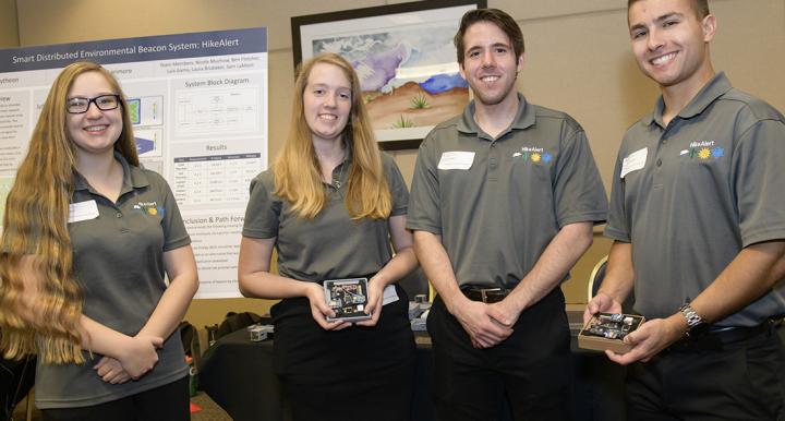 Four students in gray polo shirts stand in front of a scientific poster, holding electronic parts of a beacon system.
